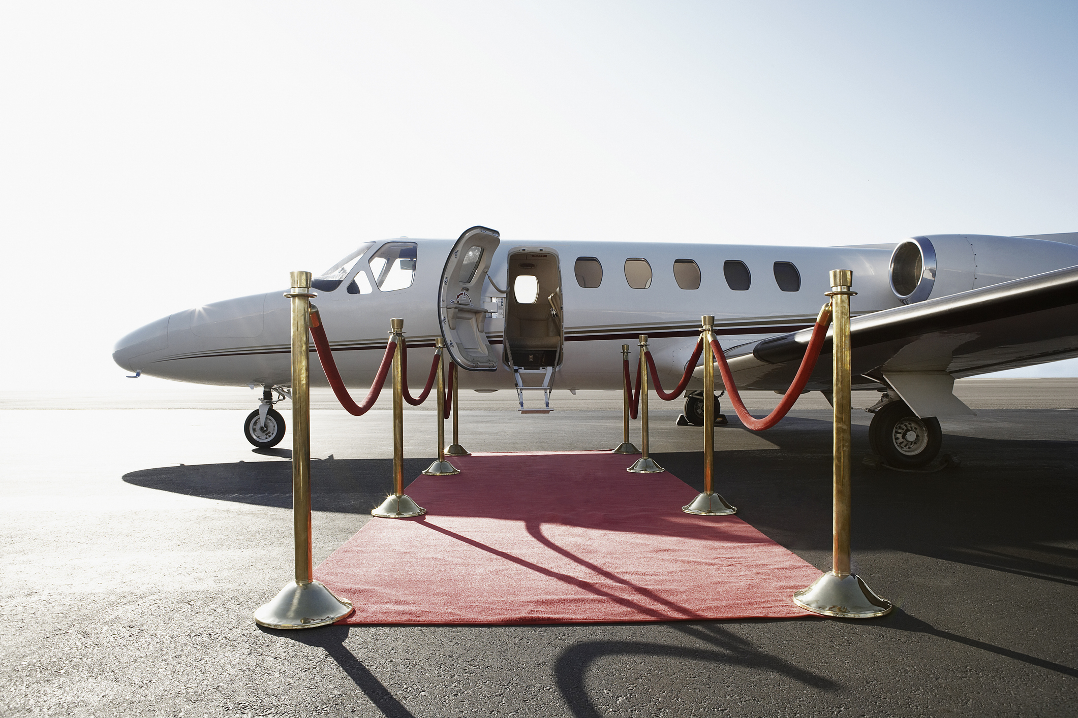 A private jet with open door is positioned at the end of a red carpet pathway lined with gold stanchions