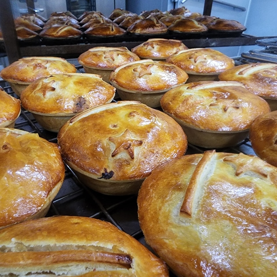 Multiple freshly baked meat pies with golden crusts, some with star-shaped designs, cooling on metal racks in a commercial kitchen