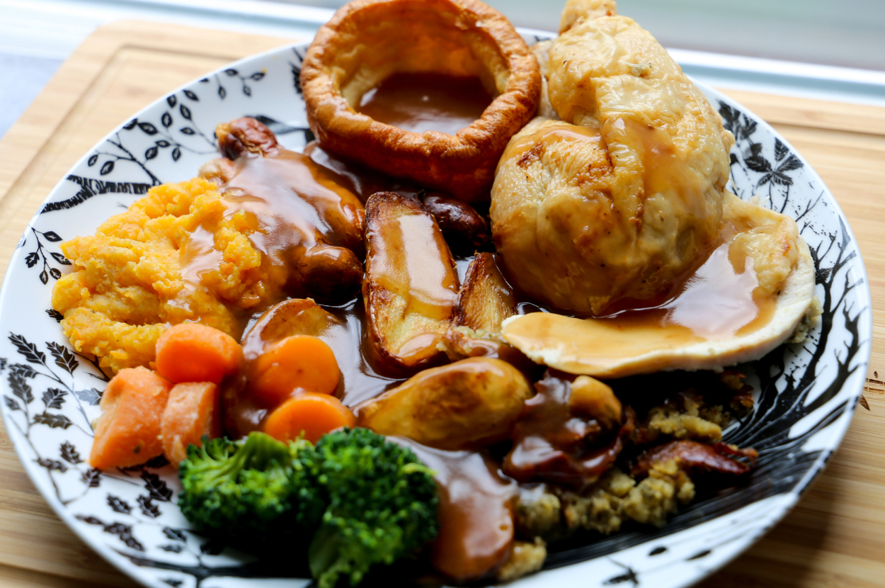 A plate of food with roast chicken, Yorkshire pudding, mashed potatoes, broccoli, carrots, and gravy