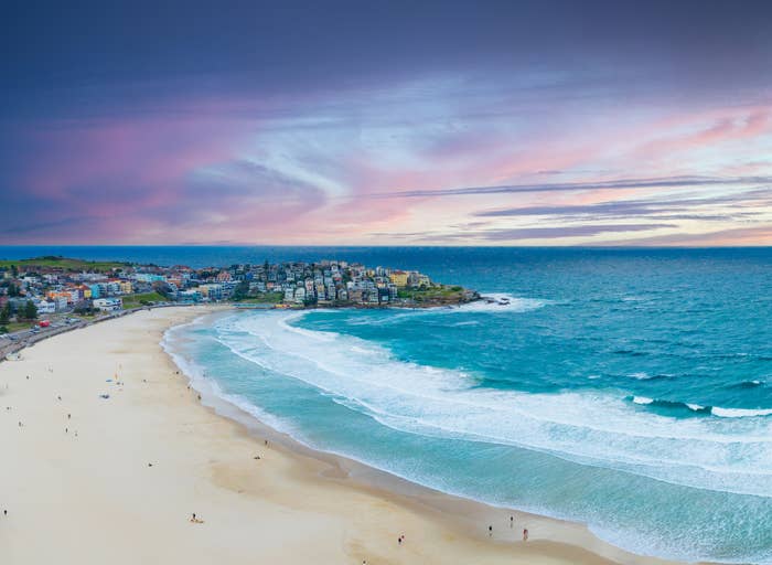 Aerial view of a beach with waves lapping the shore, a coastline lined with buildings, and a town in the background under a partly clouded sky