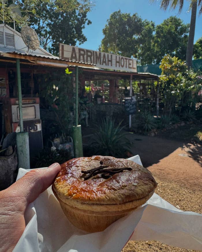 Hand holds a savory pie in front of Larrimah Hotel with greenery and rustic decor in the background