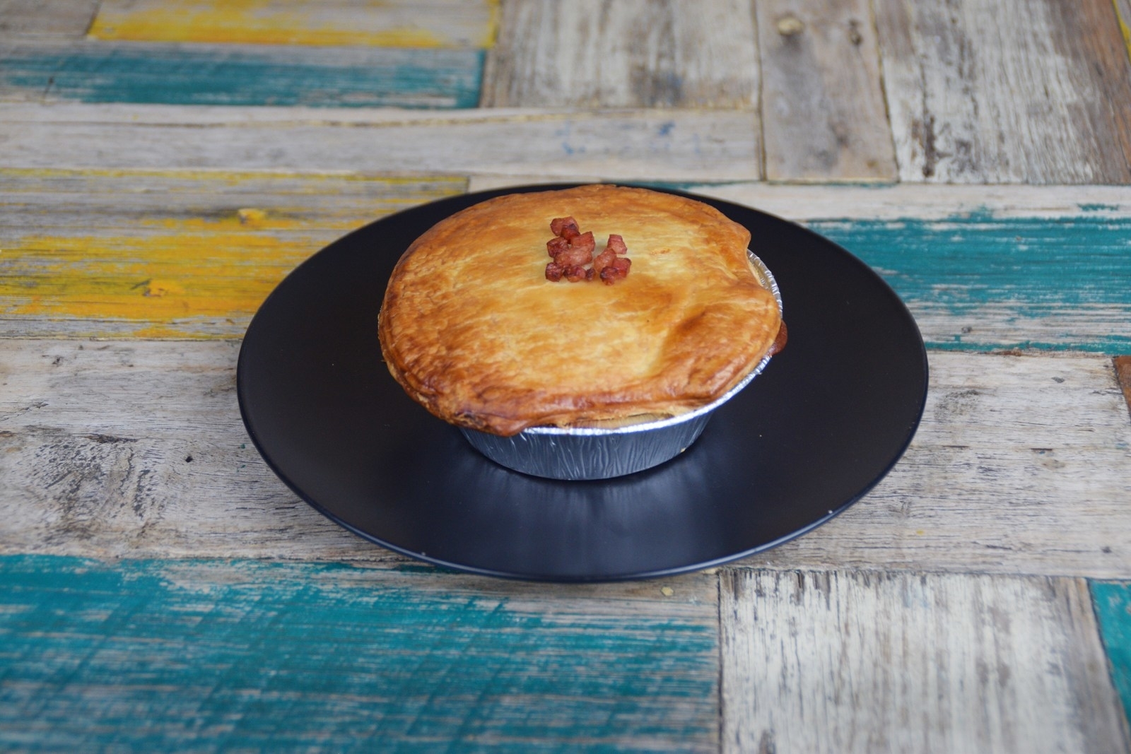A golden-brown pot pie with bacon bits on top sits on a black plate, placed on a rustic wooden table