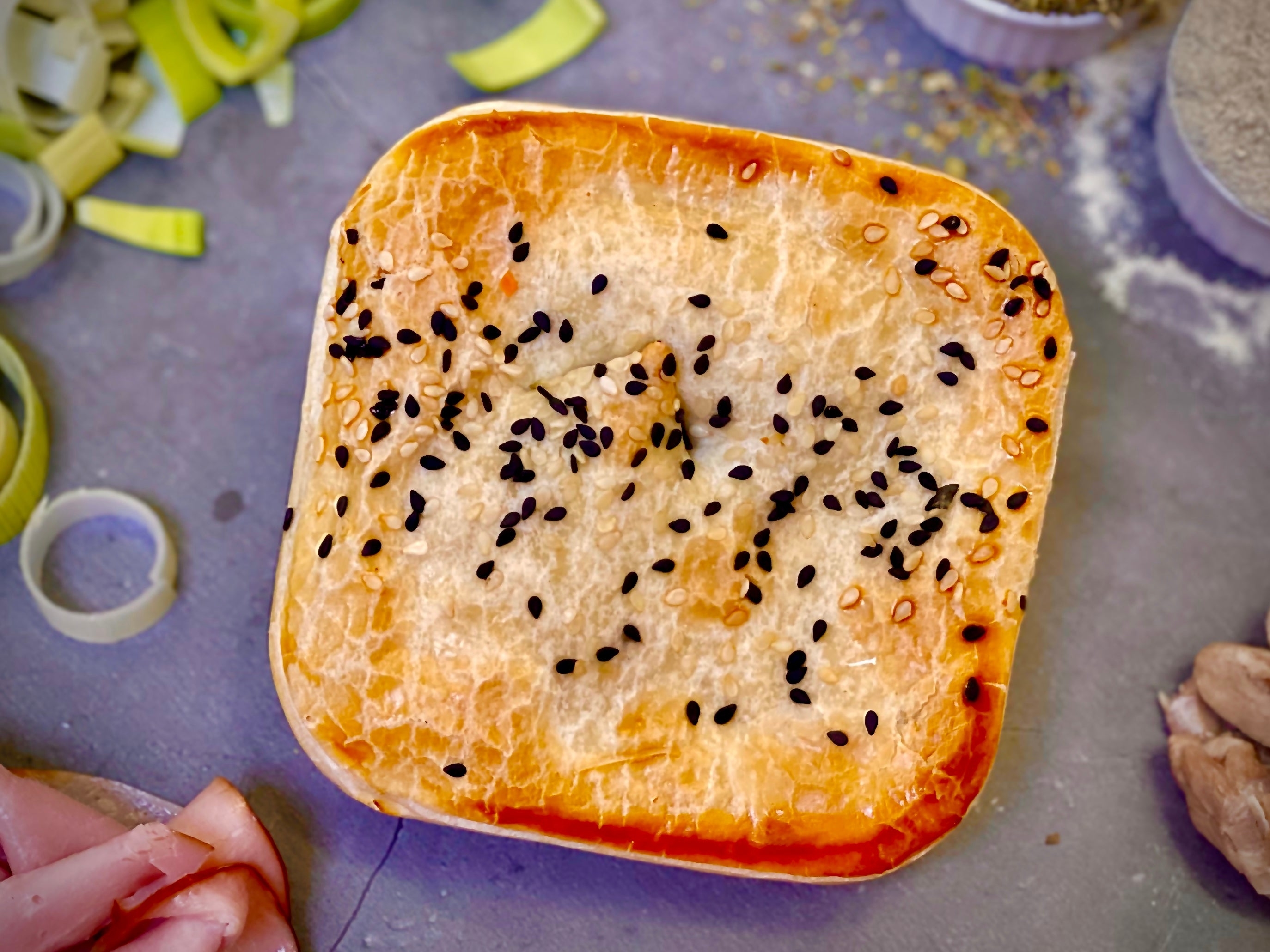 A freshly baked pastry with a golden crust, sprinkled with black sesame seeds, surrounded by chopped leeks and slices of ham on a countertop