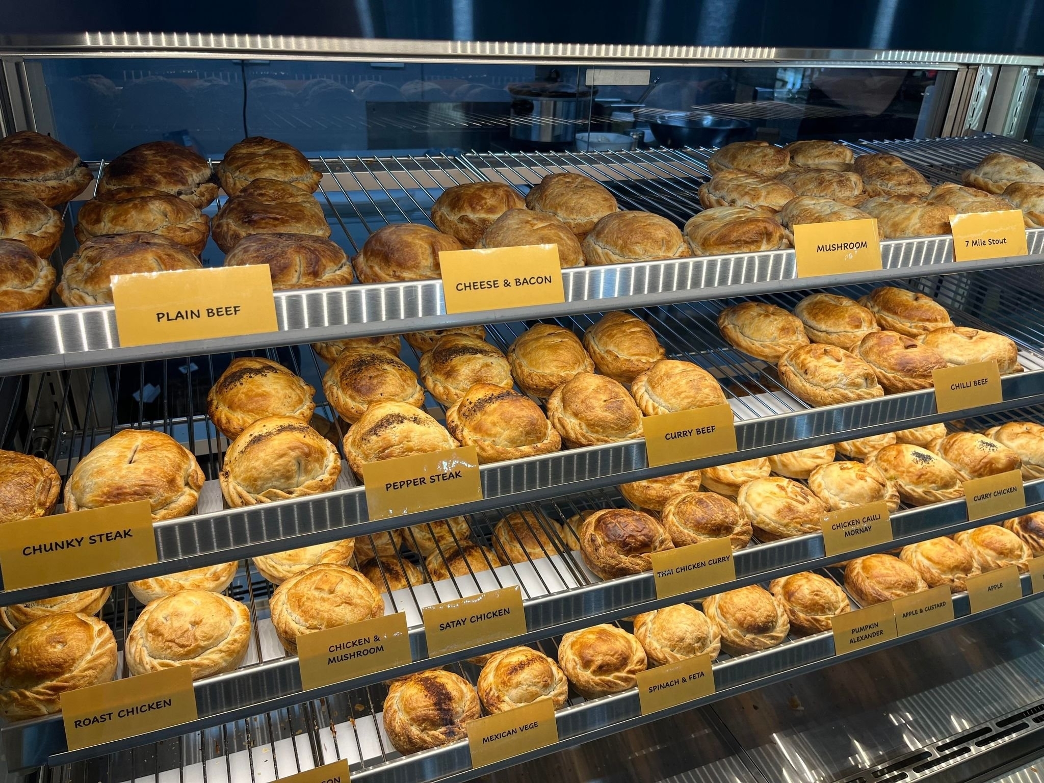 A display case filled with various pies labeled with flavors such as Plain Beef, Cheese & Bacon, Mushroom, Thai Green Curry, Pepper Steak, and Curry Beef