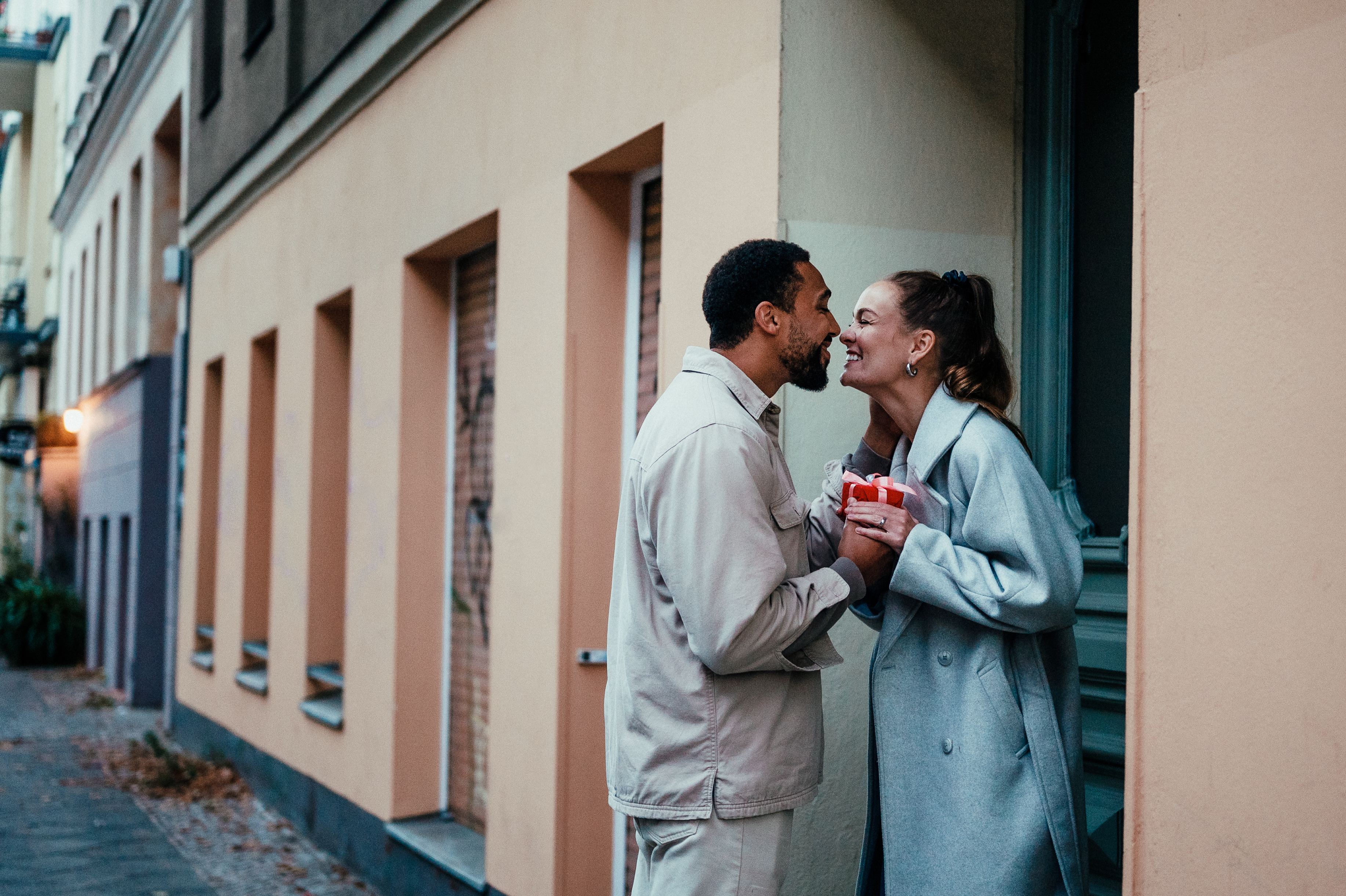 A man and woman, both smiling and holding a small gift, stand outside a building. The woman is wearing a long coat, and they appear to be laughing together
