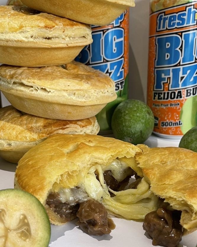 Close-up of beef and cheese pies with limes around, and two cans of FreshUp Big Fizz Feijoa Burst behind. One pie sliced open, showing the filling