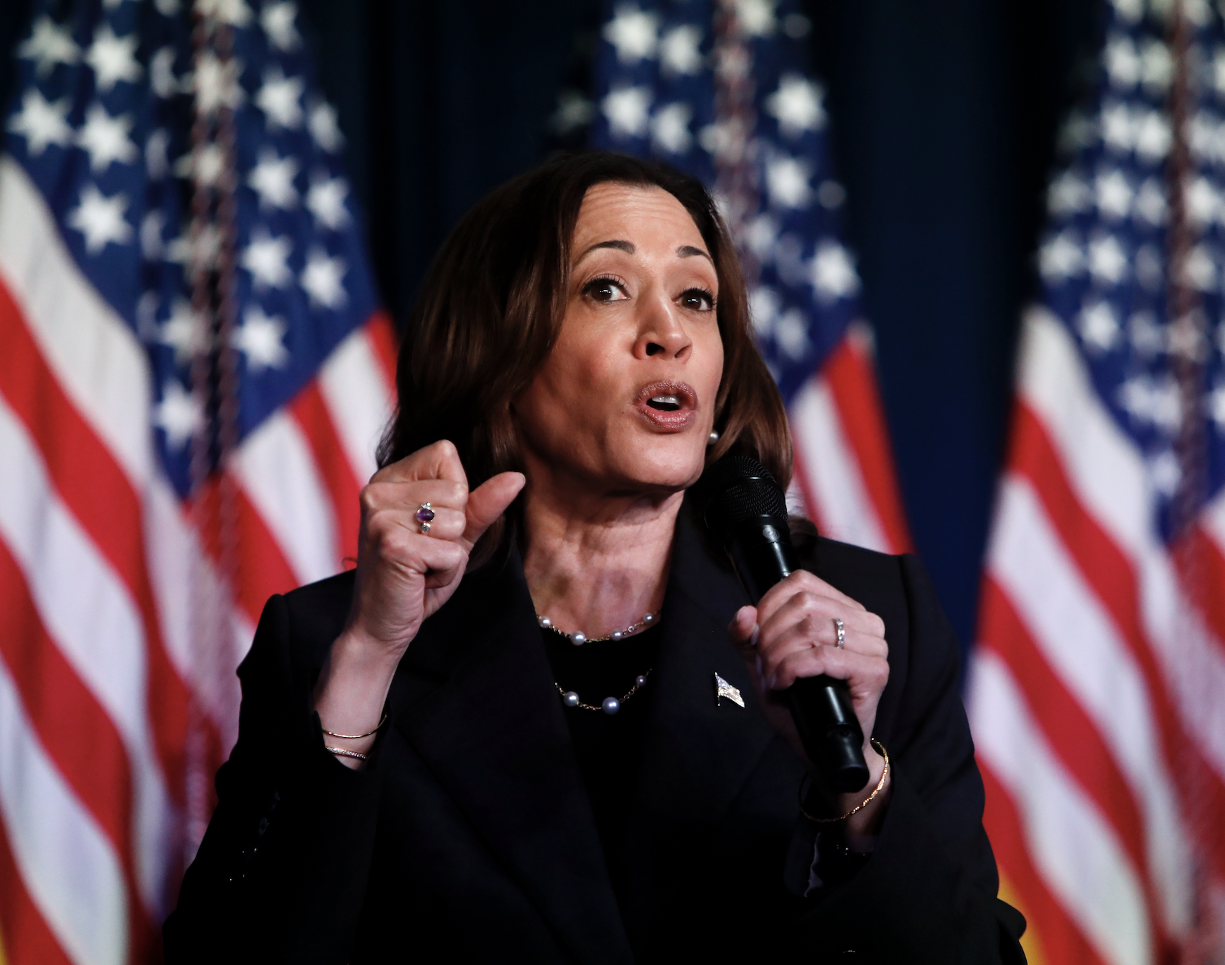 Kamala Harris speaks passionately into a microphone at an event, with multiple American flags in the background
