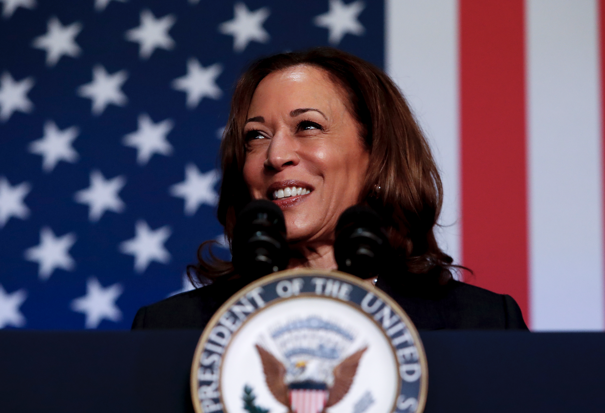 Kamala Harris speaks at a podium with a presidential seal, standing in front of an American flag