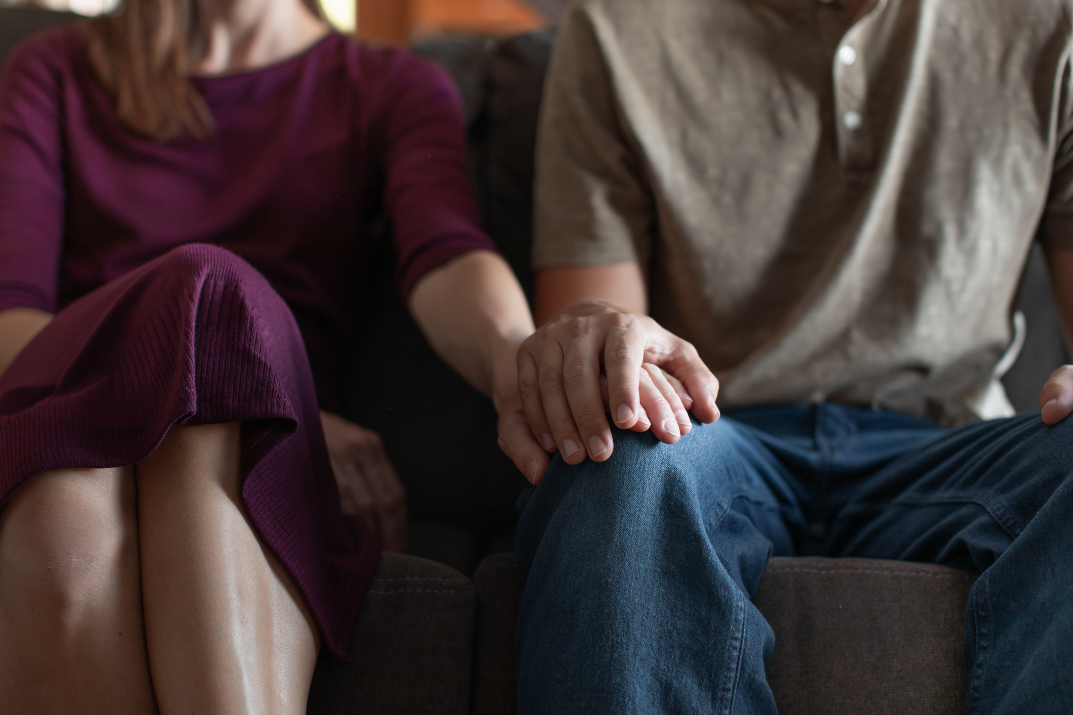 A woman and man sit on a couch, gently holding hands