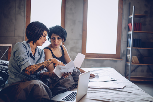 A couple sit on a bed with a laptop, analyzing documents. One person points to the papers while the other listens attentively