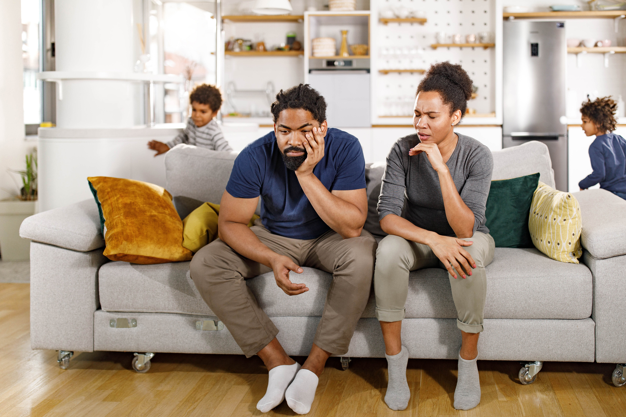 A man and woman look stressed as they sit on a couch in a living room. Two children play in the background