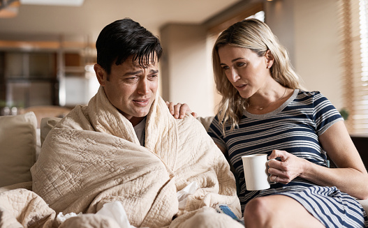 A man wrapped in a blanket sits on a couch looking distressed. A woman looks at him