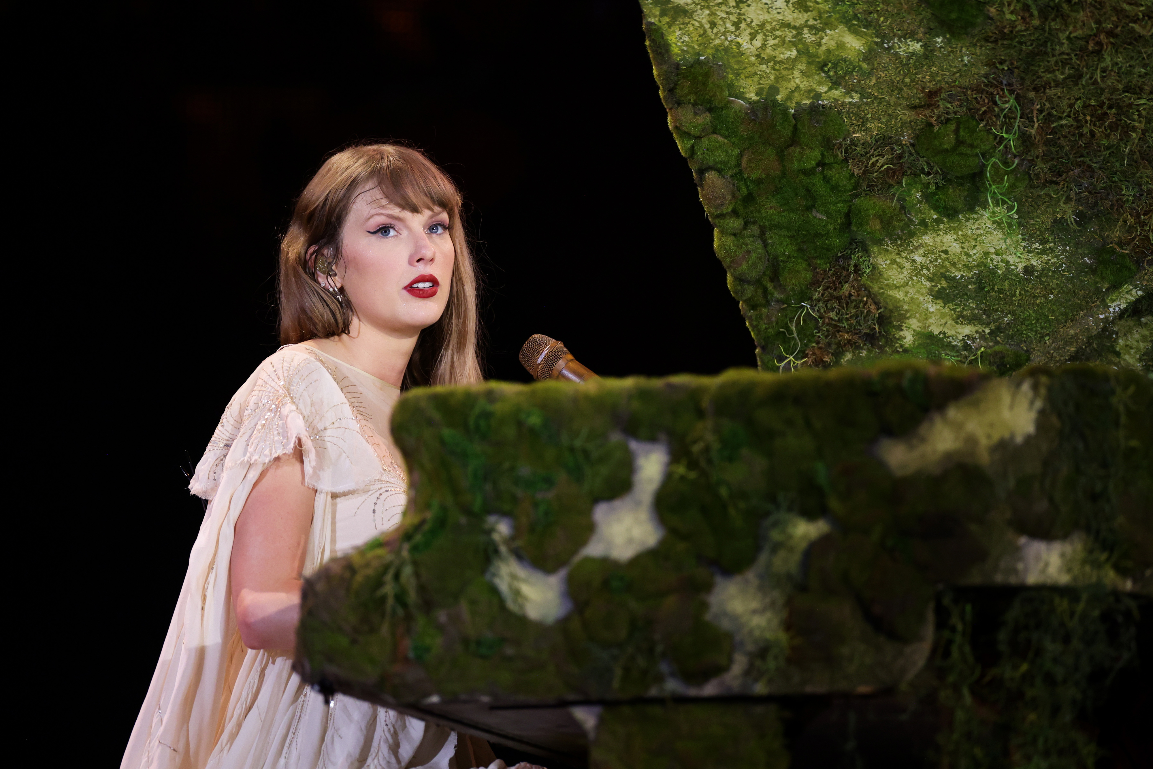 Taylor Swift in a flowing dress, seated at a green moss-covered piano, performing on stage