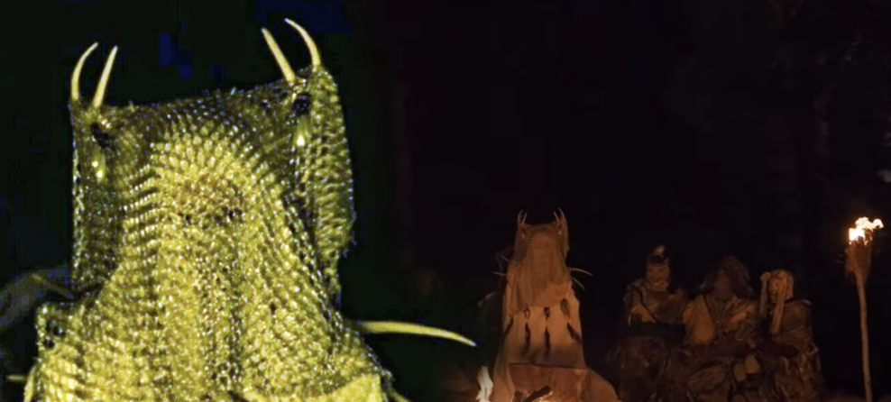 The image shows a close-up of a horned creature with scales next to a group of similarly dressed characters seated around a fire in a dark setting, likely from a TV or movie scene