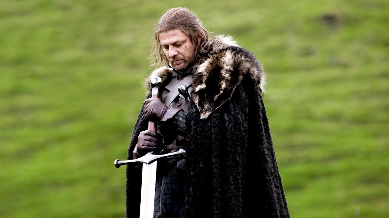 Sean Bean as Ned Stark in Game of Thrones, wearing medieval armor and a fur cloak, holding a sword, against a grassy background
