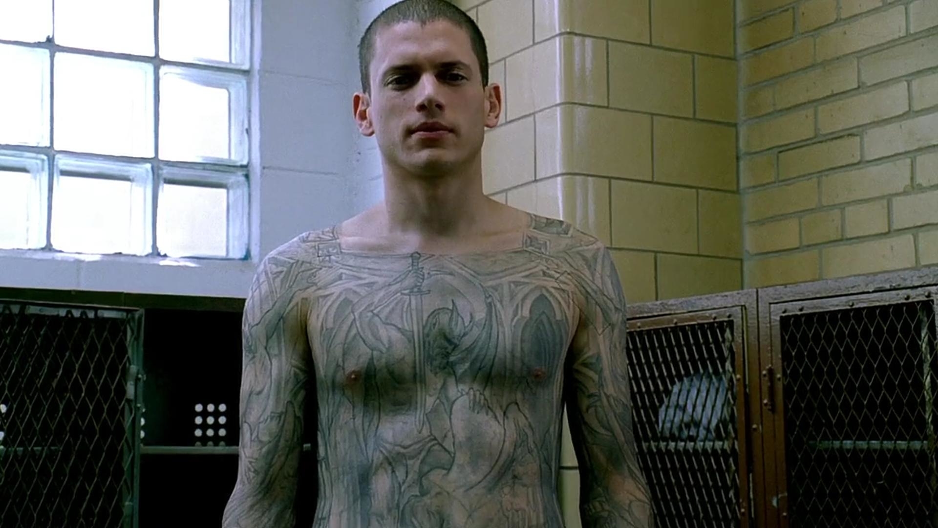Wentworth Miller stands shirtless, showcasing intricate tattoos on his upper body, in a scene from "Prison Break." He is in a room with lockers and a window
