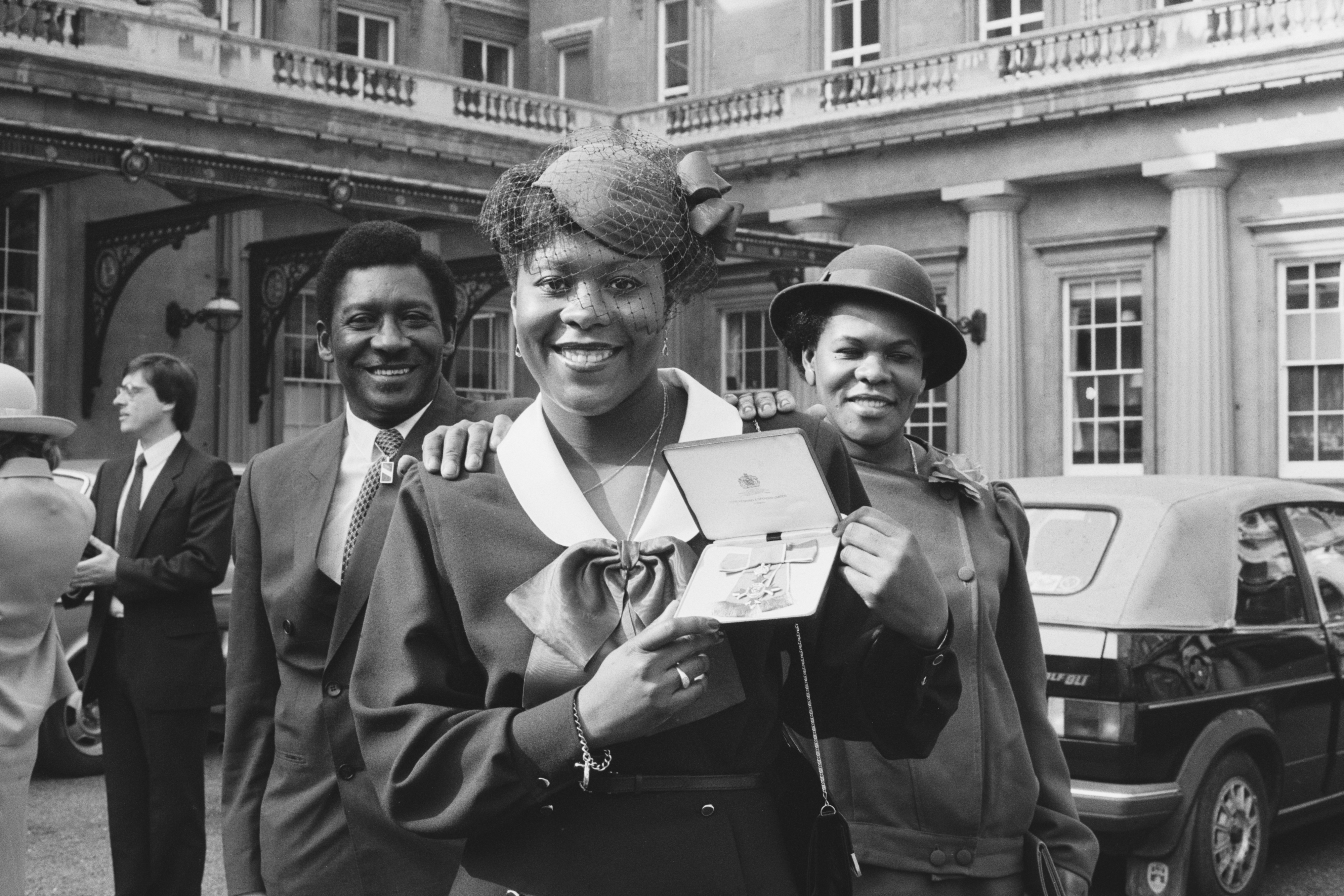 A group of three people stand outside a grand building. The person in front proudly holds up an award. Names not identified