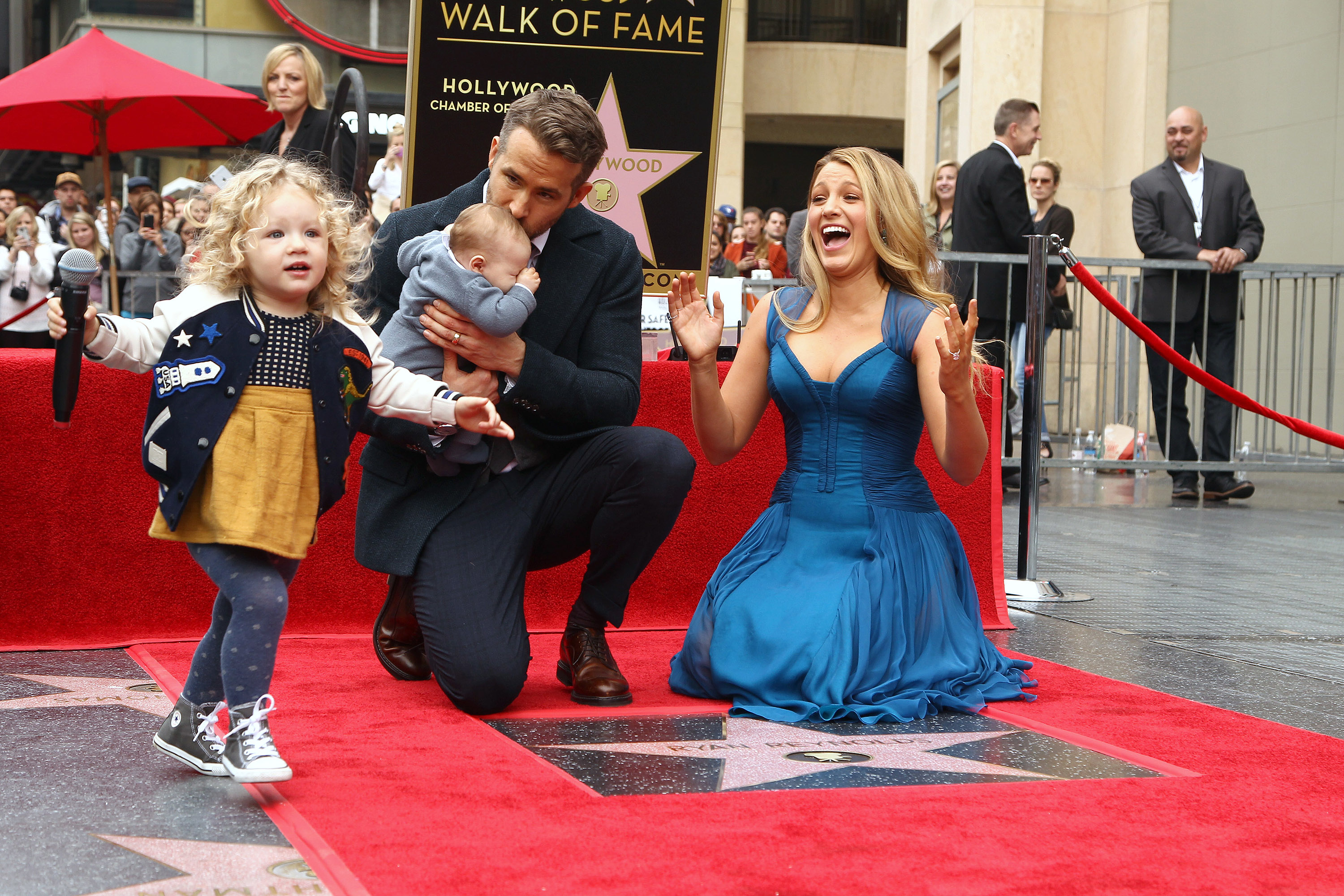 Ryan Reynolds holding a baby, Blake Lively cheering beside him, and a girl running on the Hollywood Walk of Fame star ceremony red carpet