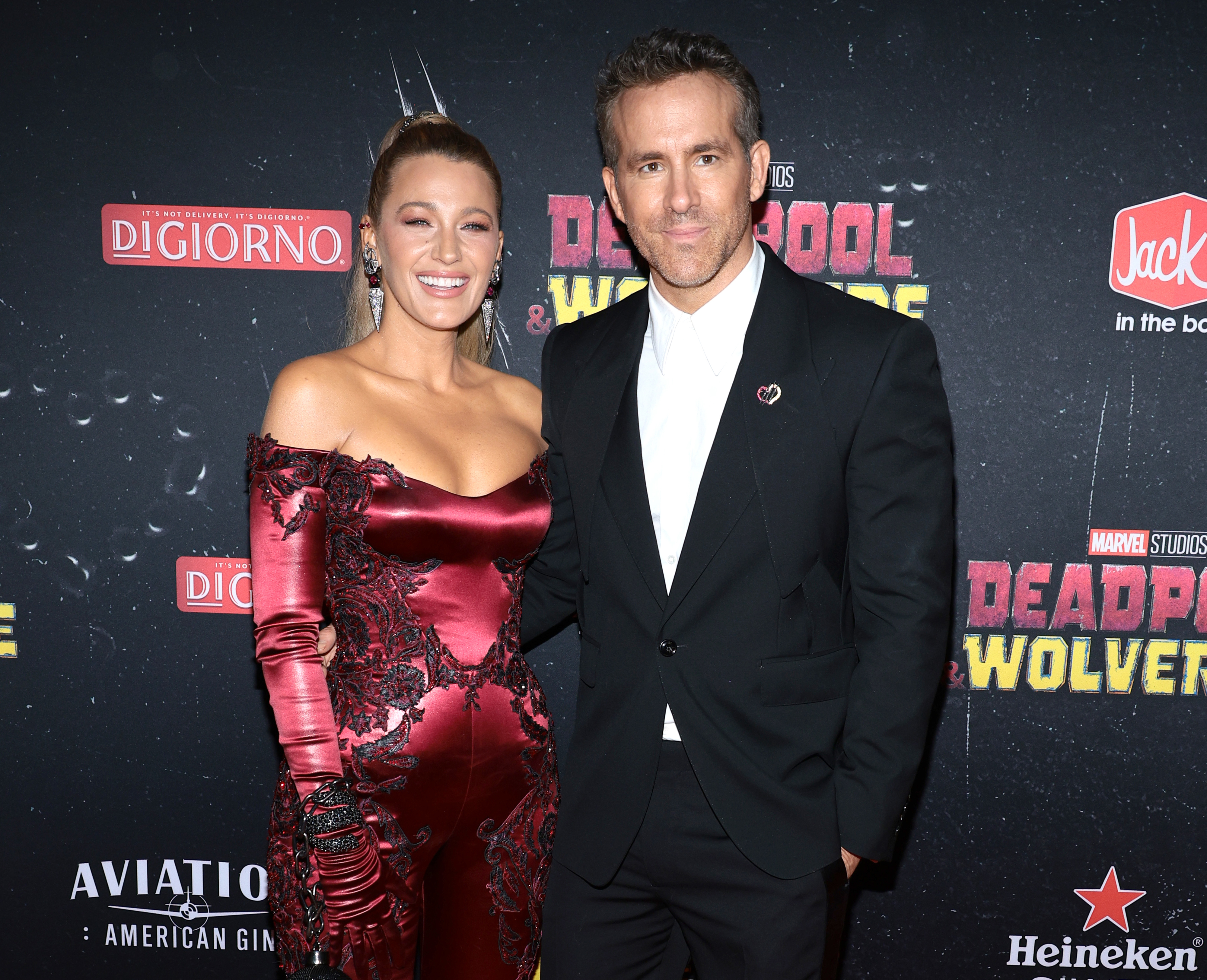 Blake Lively and Ryan Reynolds at a movie premiere. Blake is wearing an off-shoulder dress with intricate detailing, and Ryan is in a black suit with a white shirt