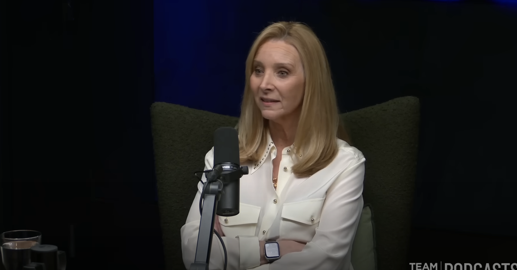 Lisa Kudrow speaking into a microphone while seated during a podcast interview. She is wearing a long-sleeve blouse
