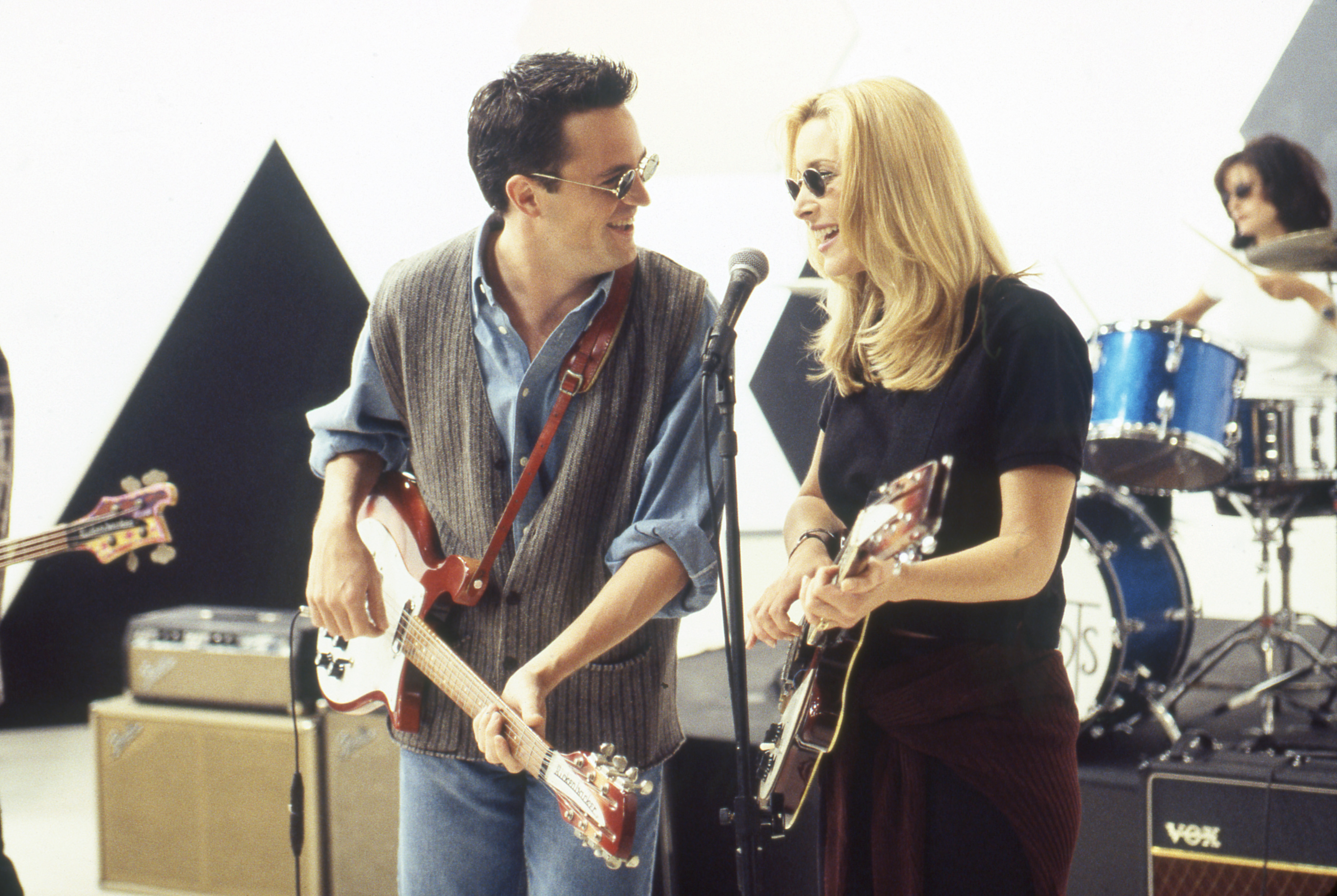 Matthew Perry and Lisa Kudrow perform on stage with guitars, wearing casual clothing. A drummer and another guitarist are playing in the background