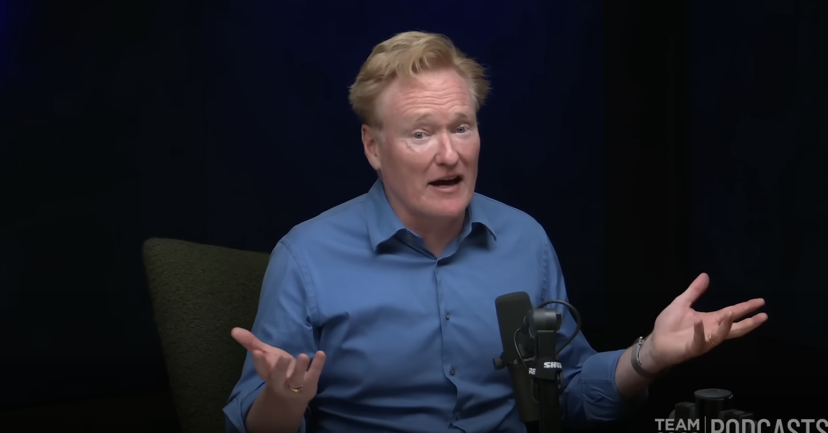 Conan O'Brien speaking into a microphone, gesturing with his hands during a podcast recording