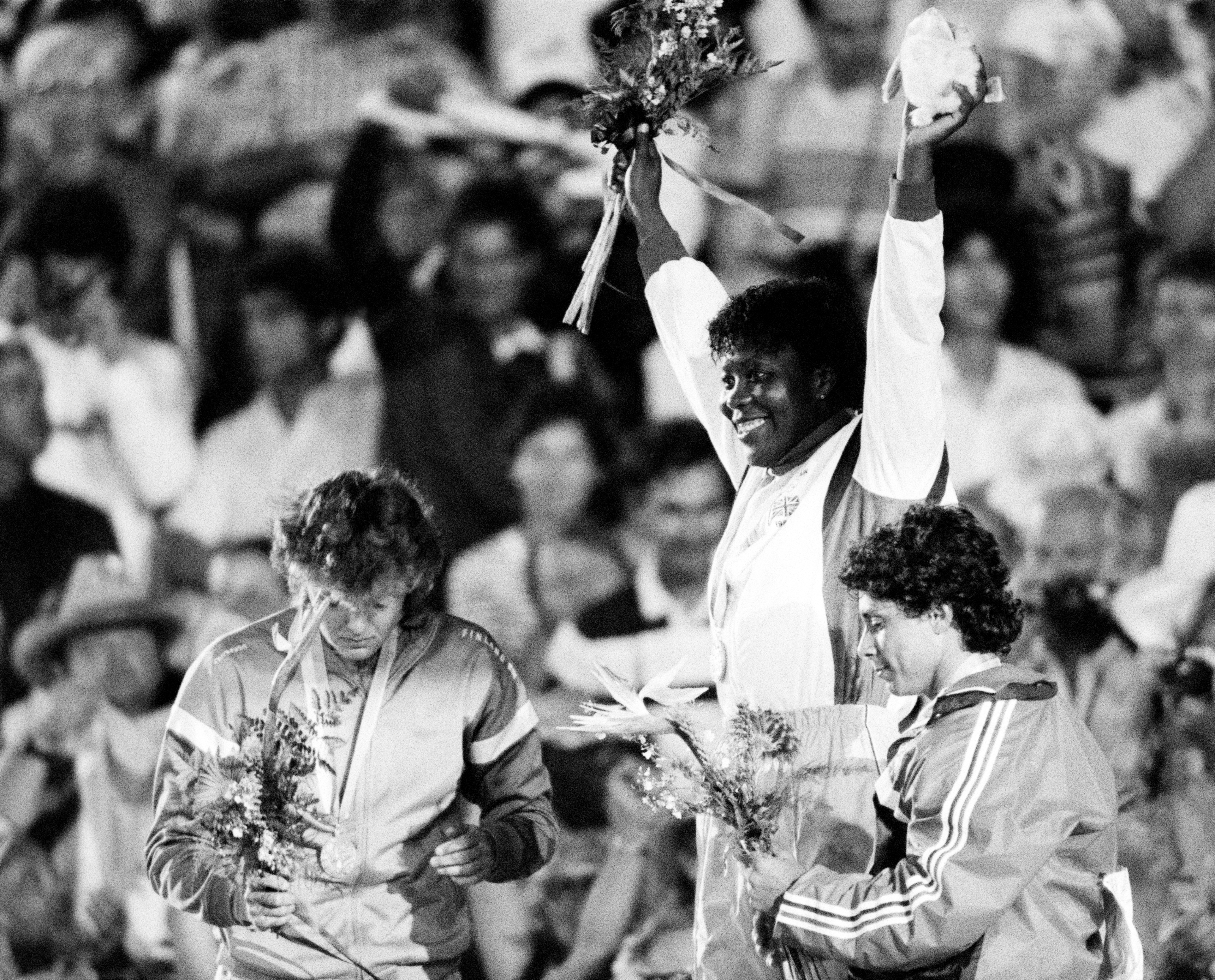 Three athletes, one on a podium holding flowers and a stuffed toy in celebration, while the other two look downward with flowers in hand, surrounded by cheering crowd