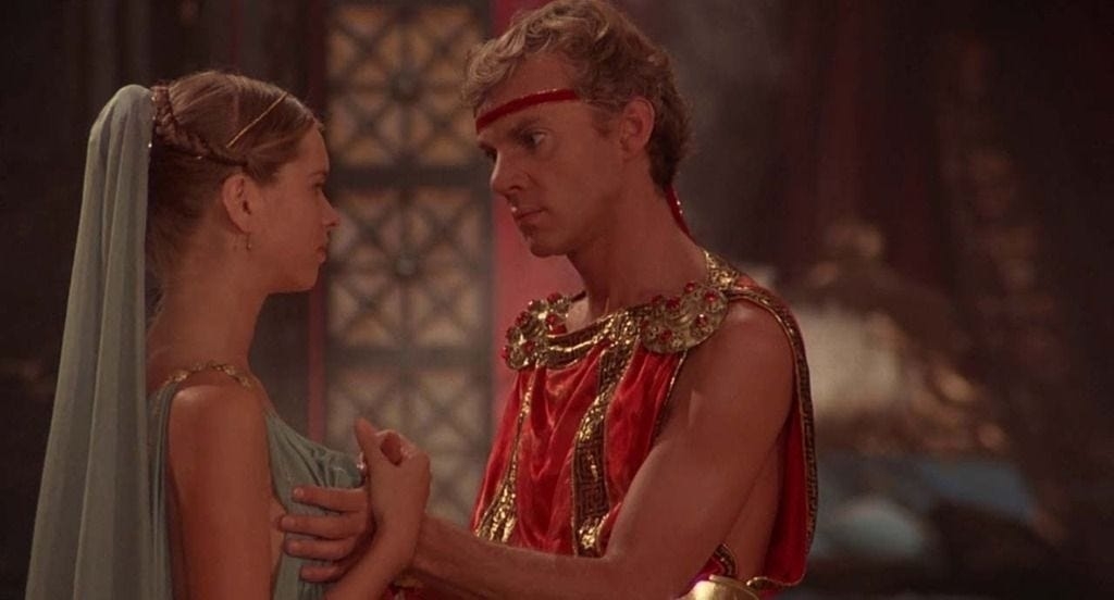 A young woman in a light dress stands closely with a man in a Roman-style outfit who gazes at her intently. The scene appears from a historical or fantasy movie