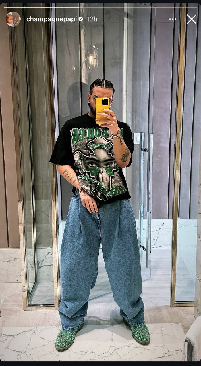 Drake takes a mirror selfie, wearing a football graphic T-shirt, baggy jeans, and green shoes