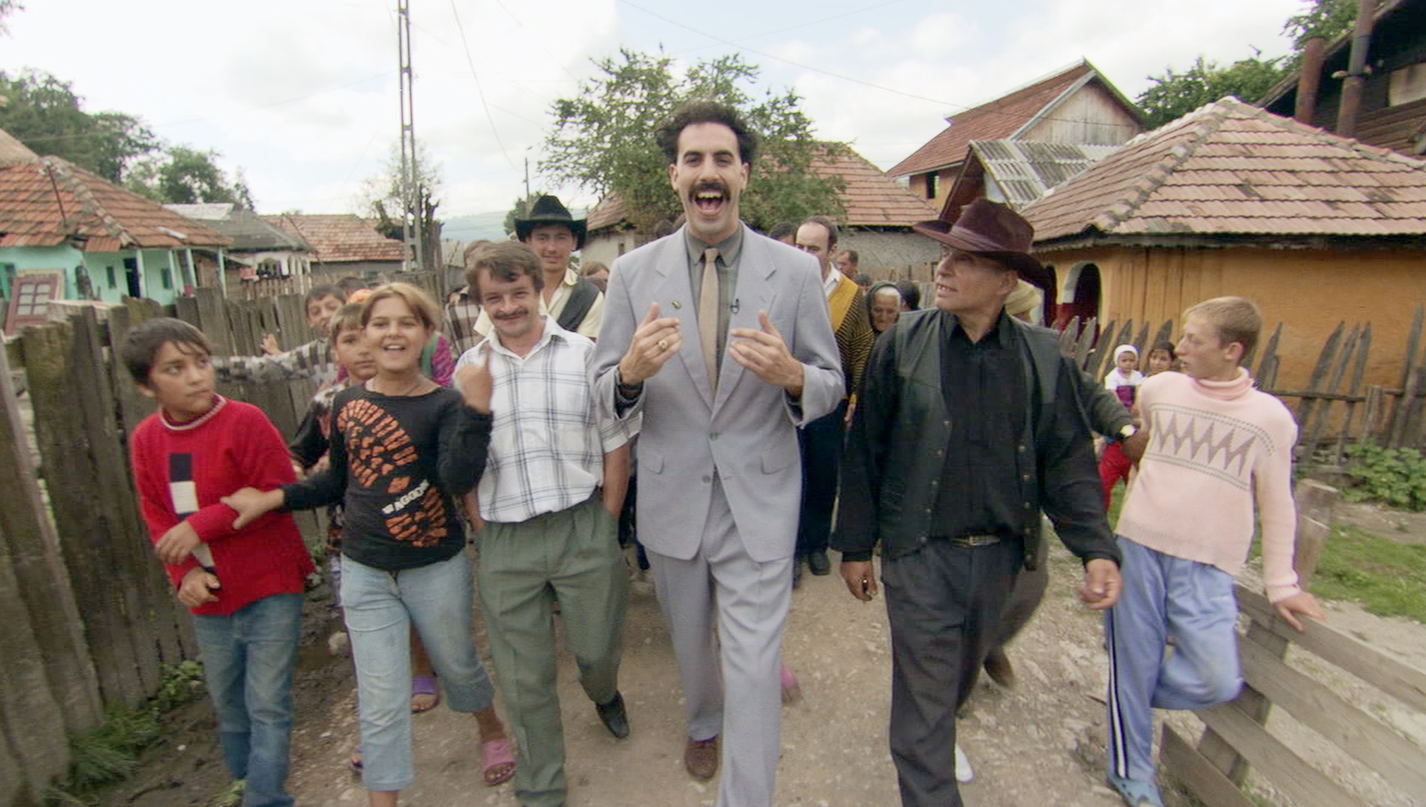 Sacha Baron Cohen as Borat, dressed in a light suit, walks through a village joyfully surrounded by townspeople. Scene from the film "Borat."