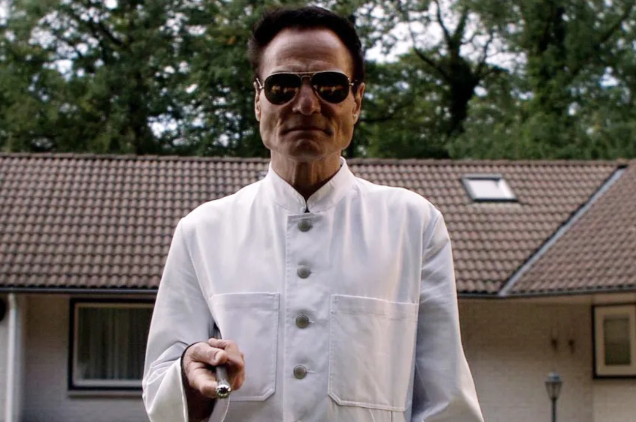 A person in a white buttoned coat and dark sunglasses stands in front of a house holding a pen-like object, under a backdrop of trees