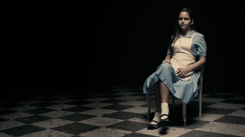 A young girl dressed as Alice from Alice in Wonderland sits alone on a chair in a dimly lit room with a checkered floor