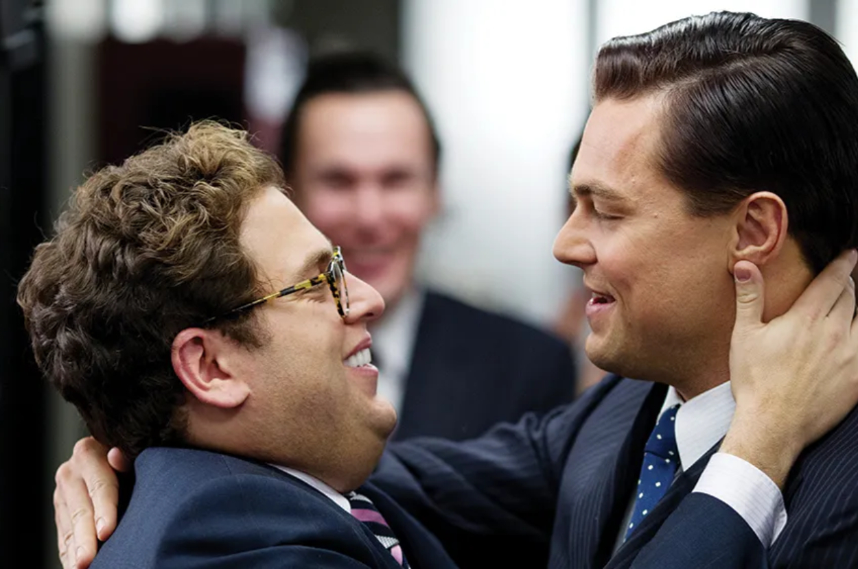 Leonardo DiCaprio and Jonah Hill in suits, smiling and embracing, with a blurred person in the background, likely from a scene in a movie