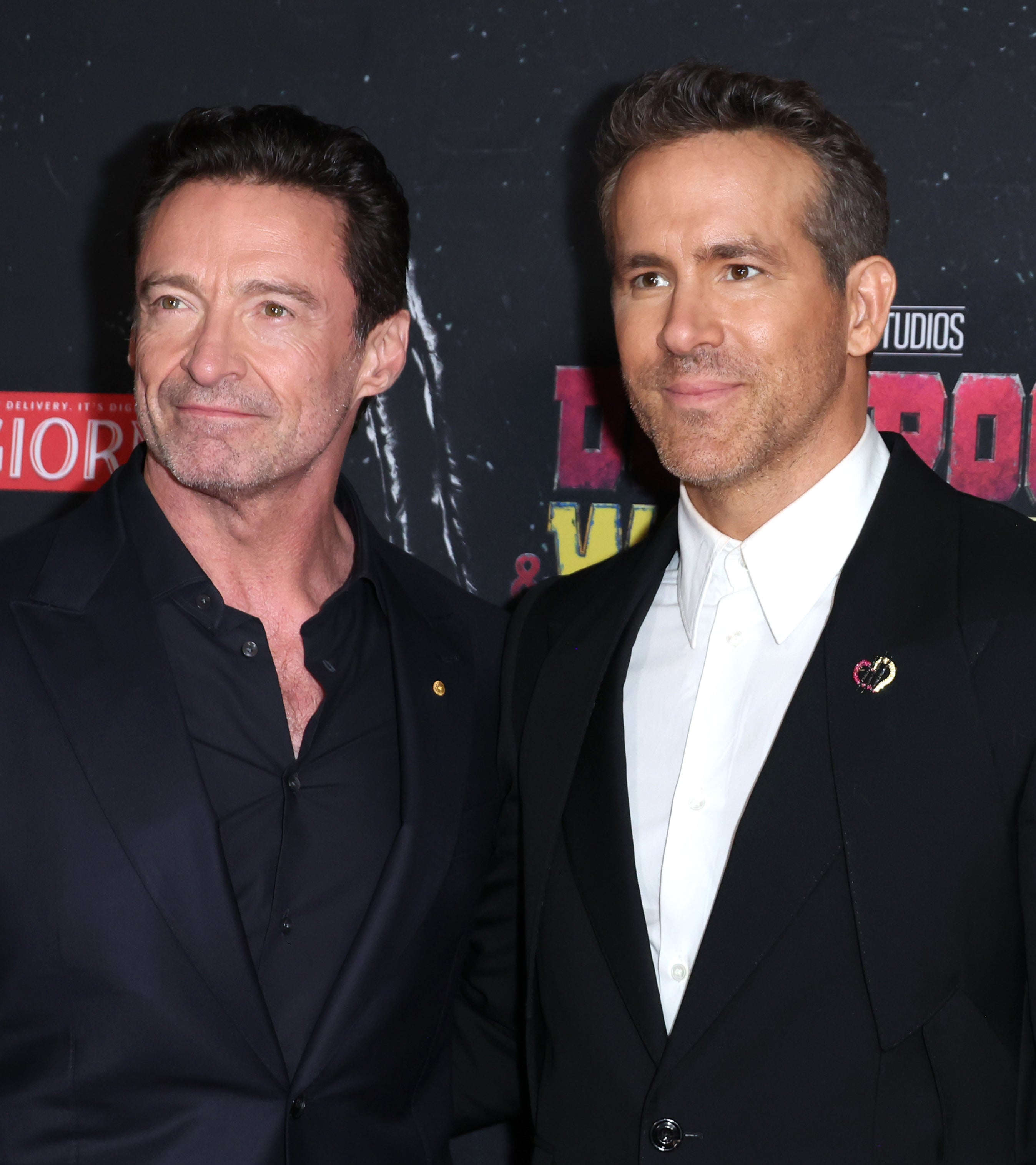 Hugh Jackman and Ryan Reynolds on the red carpet, both wearing black suits, at the Deadpool and Wolverine event