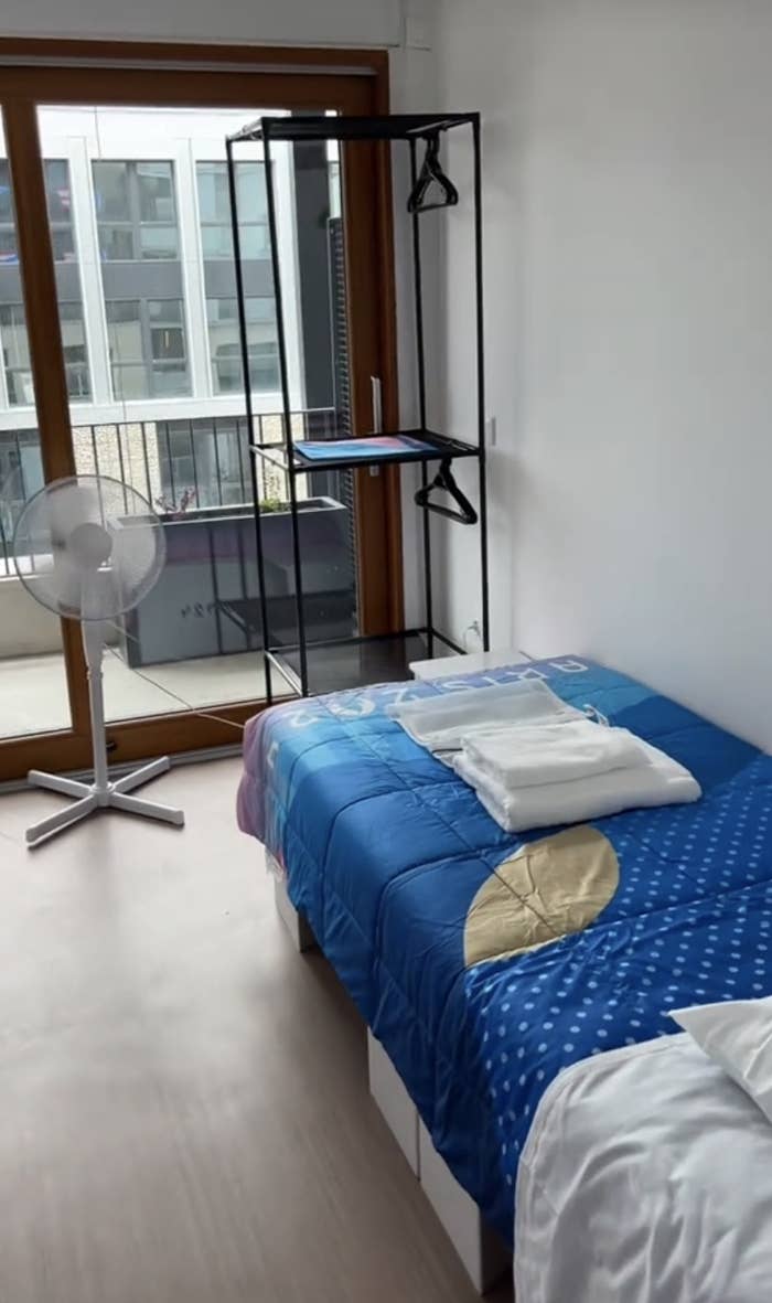 A modern bedroom with a bed, neatly folded linens on top, a standing fan, and an empty shelving unit near a balcony with large windows