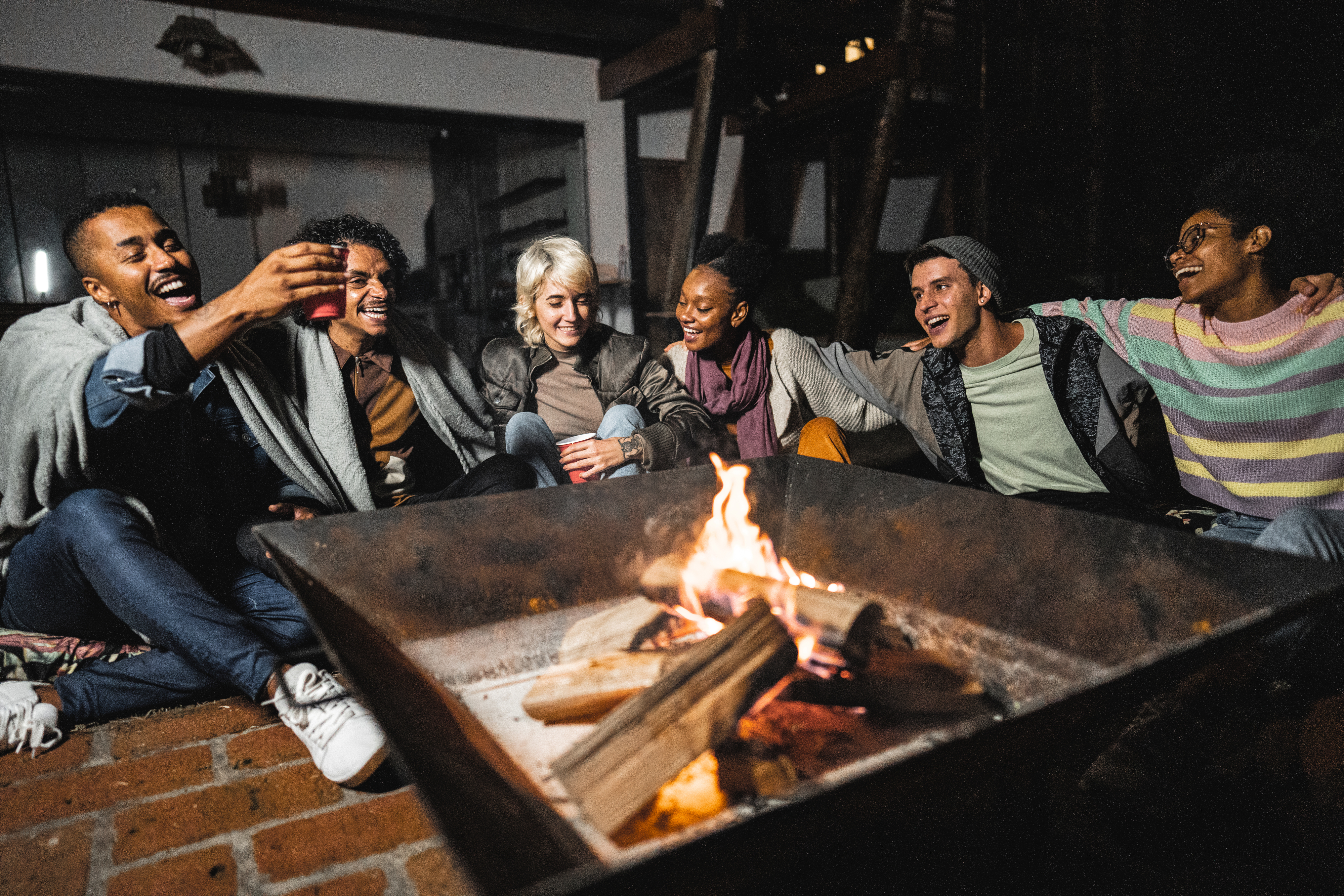 Group of six people sitting around a fire pit, laughing and toasting with drinks