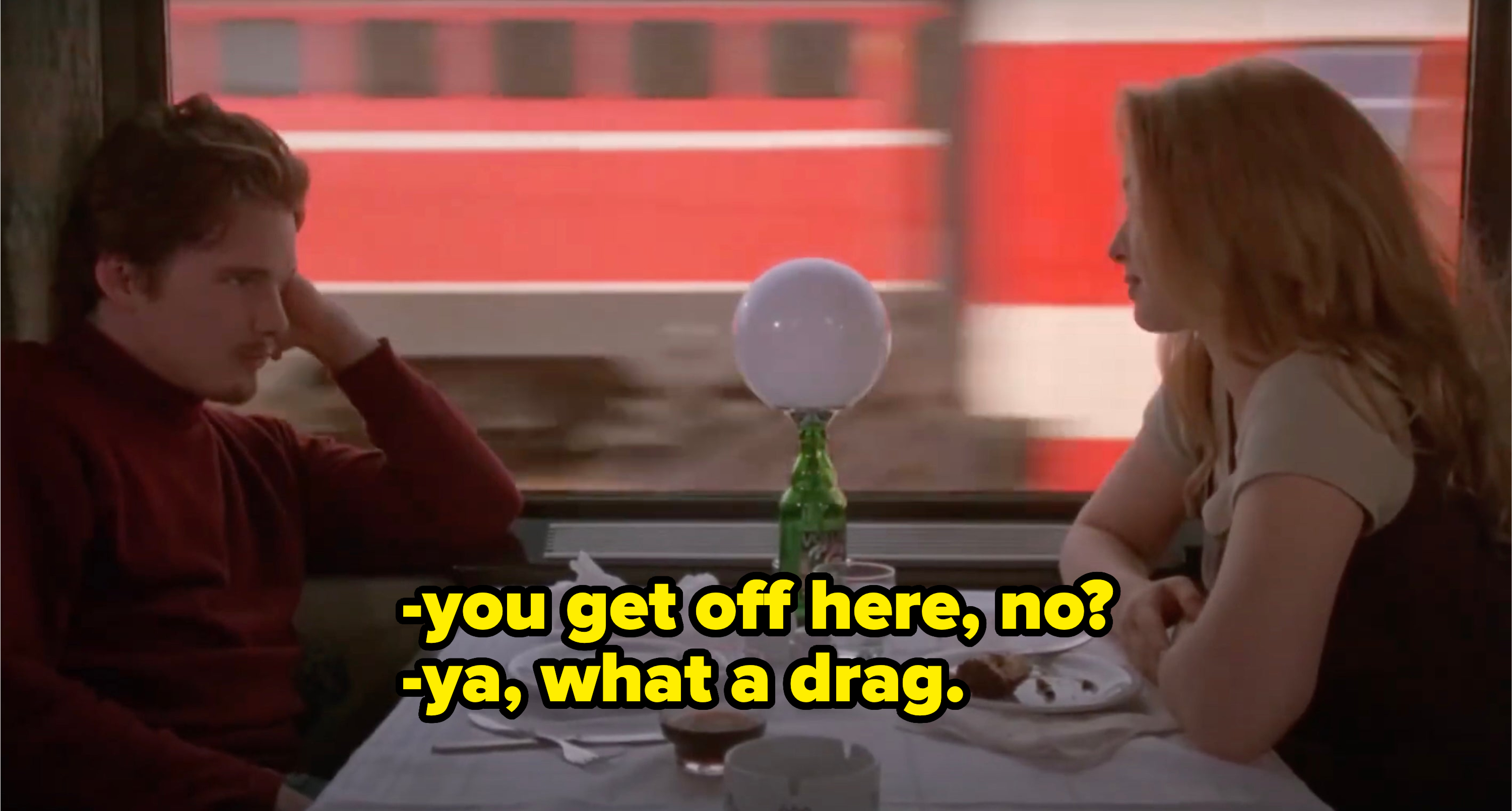 Ethan Hawke and Julie Delpy sit across each other at a table on a train, with a red train passing by outside the window