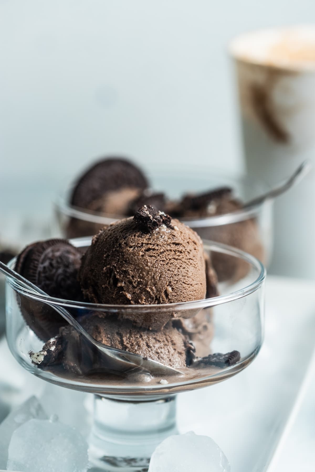 Two glass bowls filled with scoops of chocolate ice cream, topped with Oreo cookies, are placed on a tray. A cup of coffee is blurred in the background