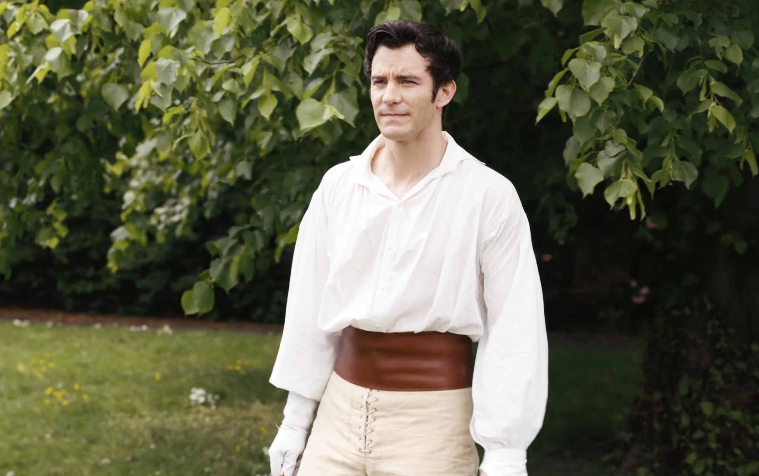 Luke Thompson, in a scene, wearing a loose shirt tucked into high-waisted trousers with a wide belt, standing outdoors with trees in the background