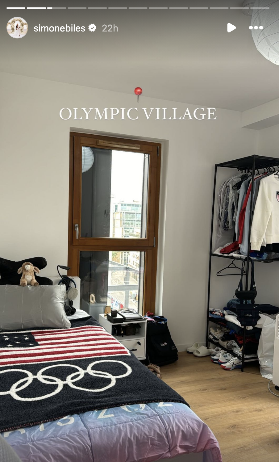 Simone Biles' Instagram Story shows a tidy room in the Olympic Village, featuring a bed with an Olympic-themed blanket and a wardrobe beside a window