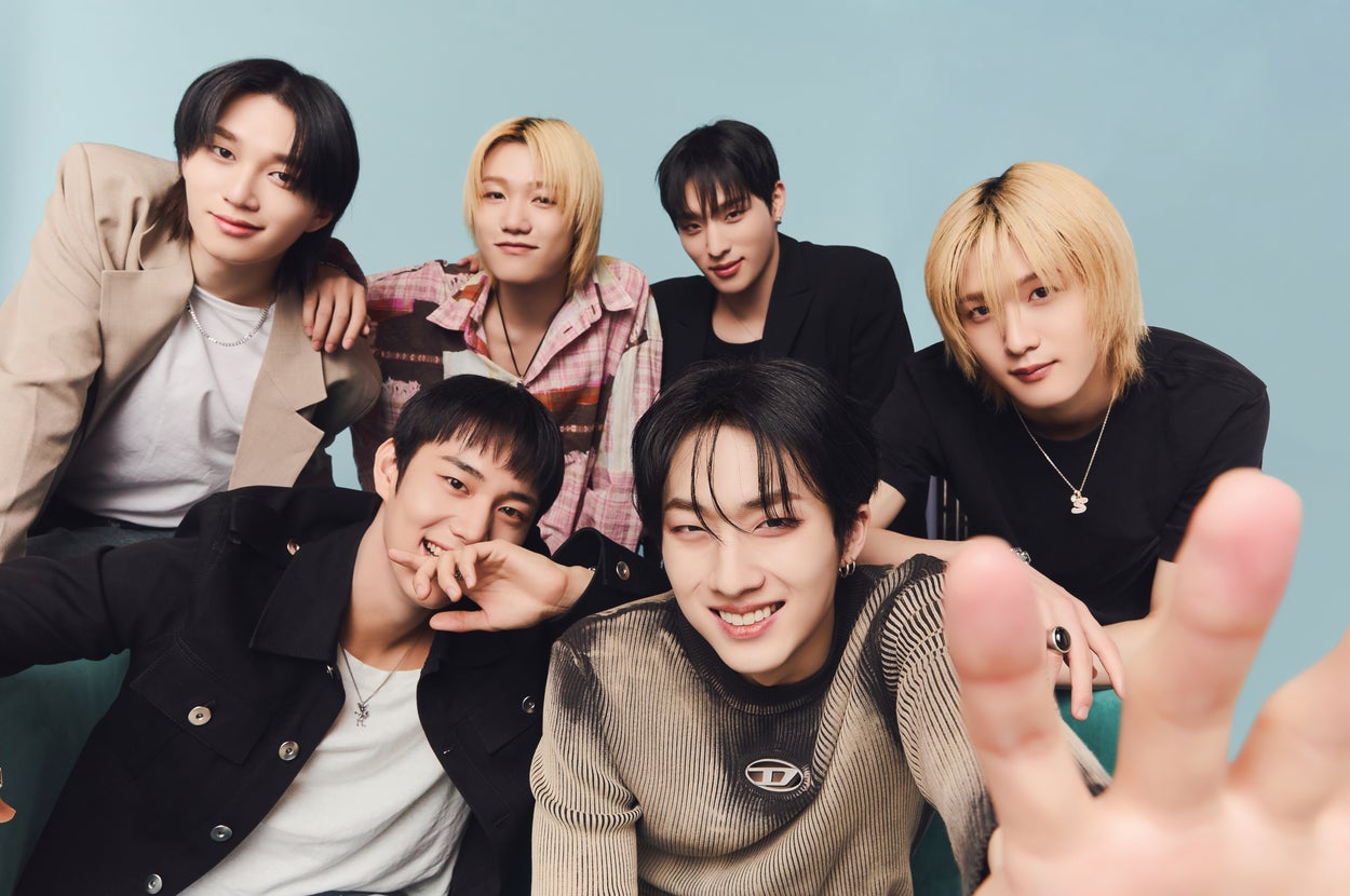 Six members of the K-pop group ENHYPEN posing together in fashionable attire, smiling and making peace signs towards the camera