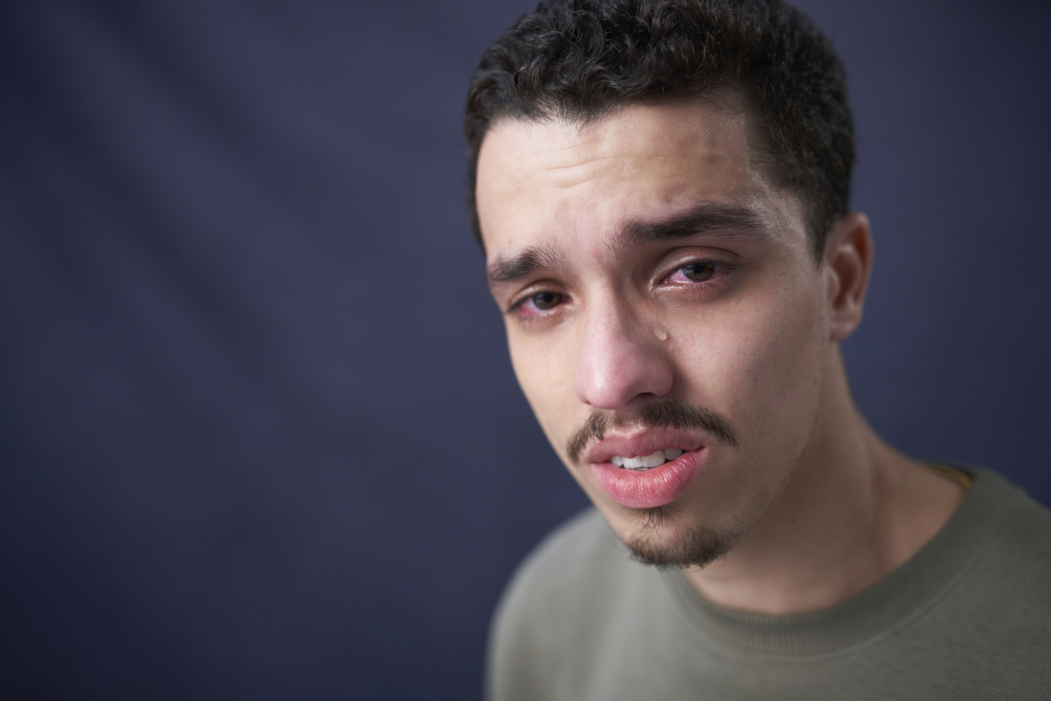 A person with short curly hair and a mustache appears emotional, with a tear on their cheek. The person is wearing a plain shirt against a plain background
