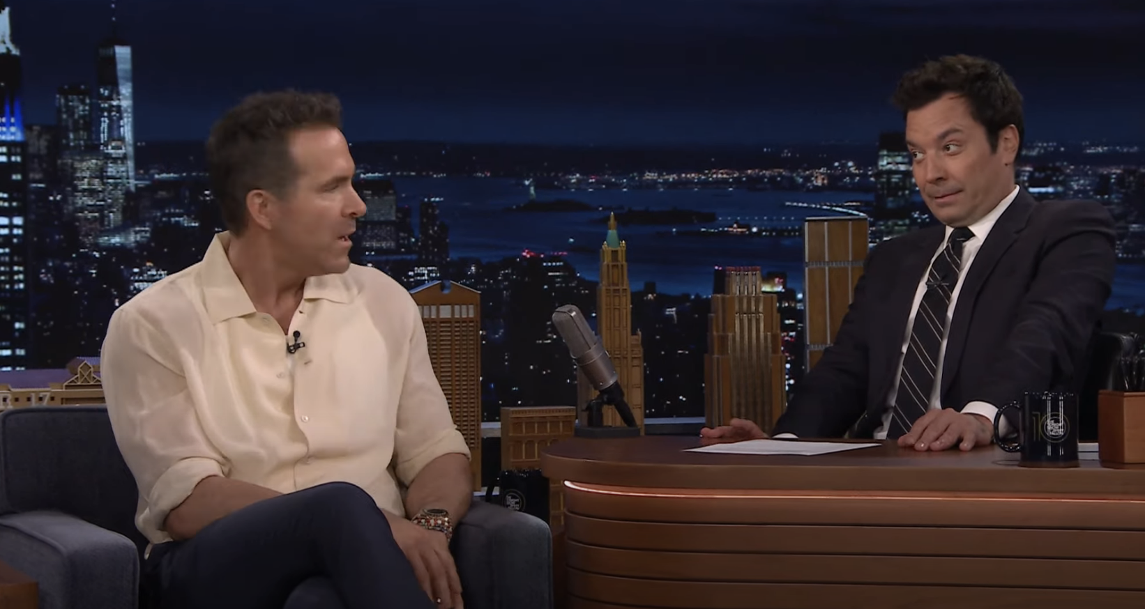 Ryan Reynolds, in a casual shirt and dark pants, talks to Jimmy Fallon, in a suit and tie, on &quot;The Tonight Show&quot; set with a cityscape background