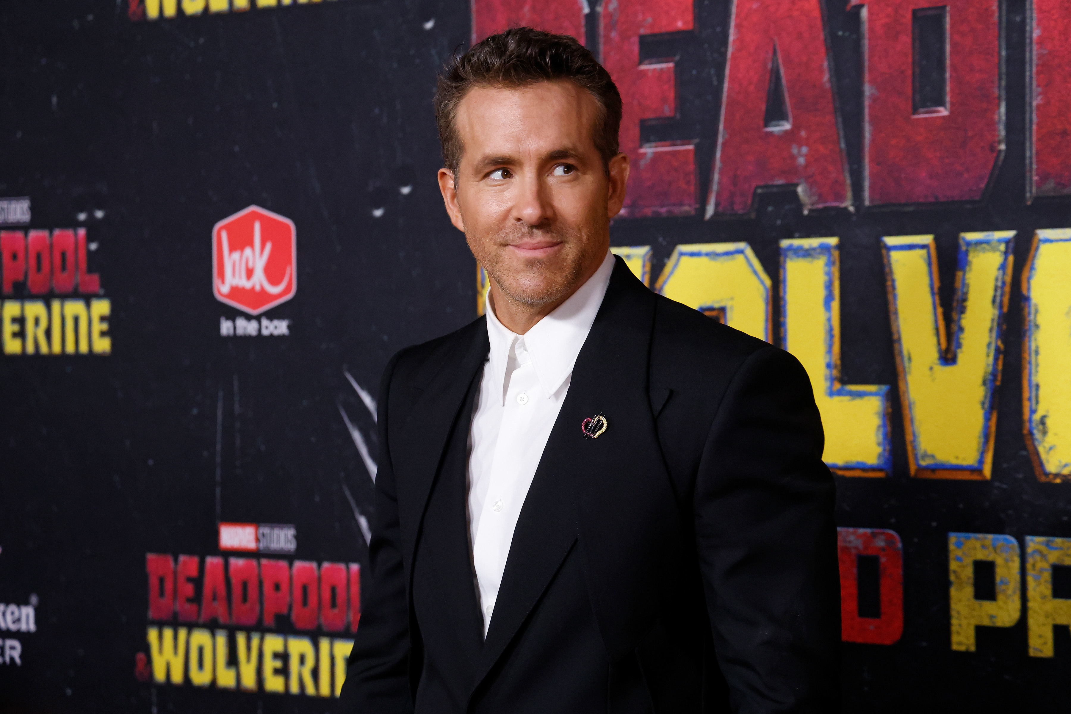 Ryan Reynolds stands in front of a &quot;Deadpool 3&quot; and &quot;Wolverine&quot; promotional backdrop, wearing a black suit with a white shirt