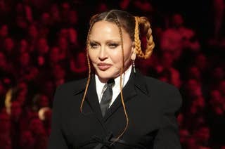 Madonna at a public event wearing a formal black suit with a white shirt and black tie. Her hair is styled in braided updos with long braided strands framing her face
