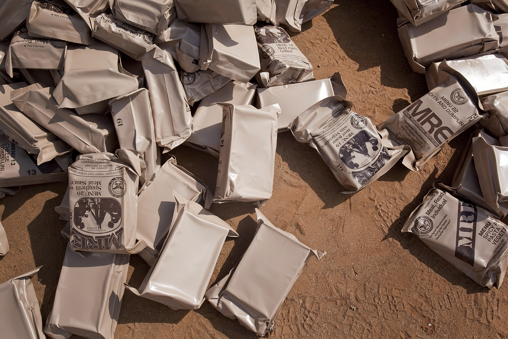 Packets of MRE (Meals Ready-to-Eat) are spread out on a dirt surface, likely at a distribution site