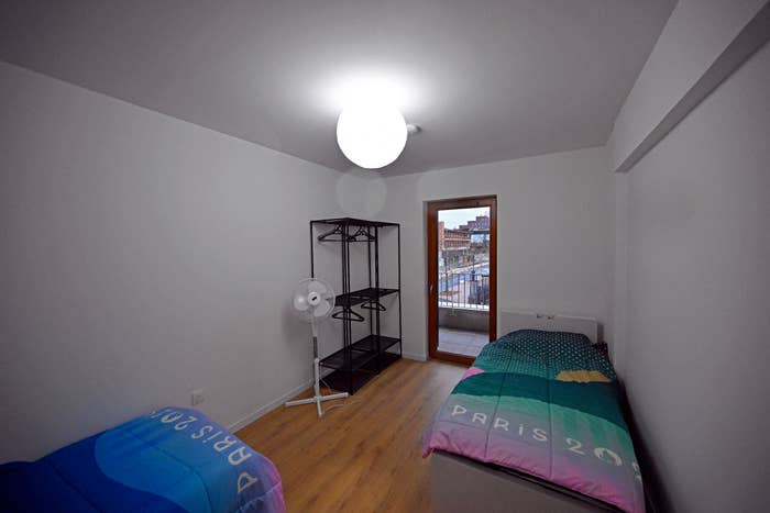 A bedroom with two single beds covered with Paris 2024 Olympic Games blankets, a standing fan, and a shelf. There is a balcony door leading outside