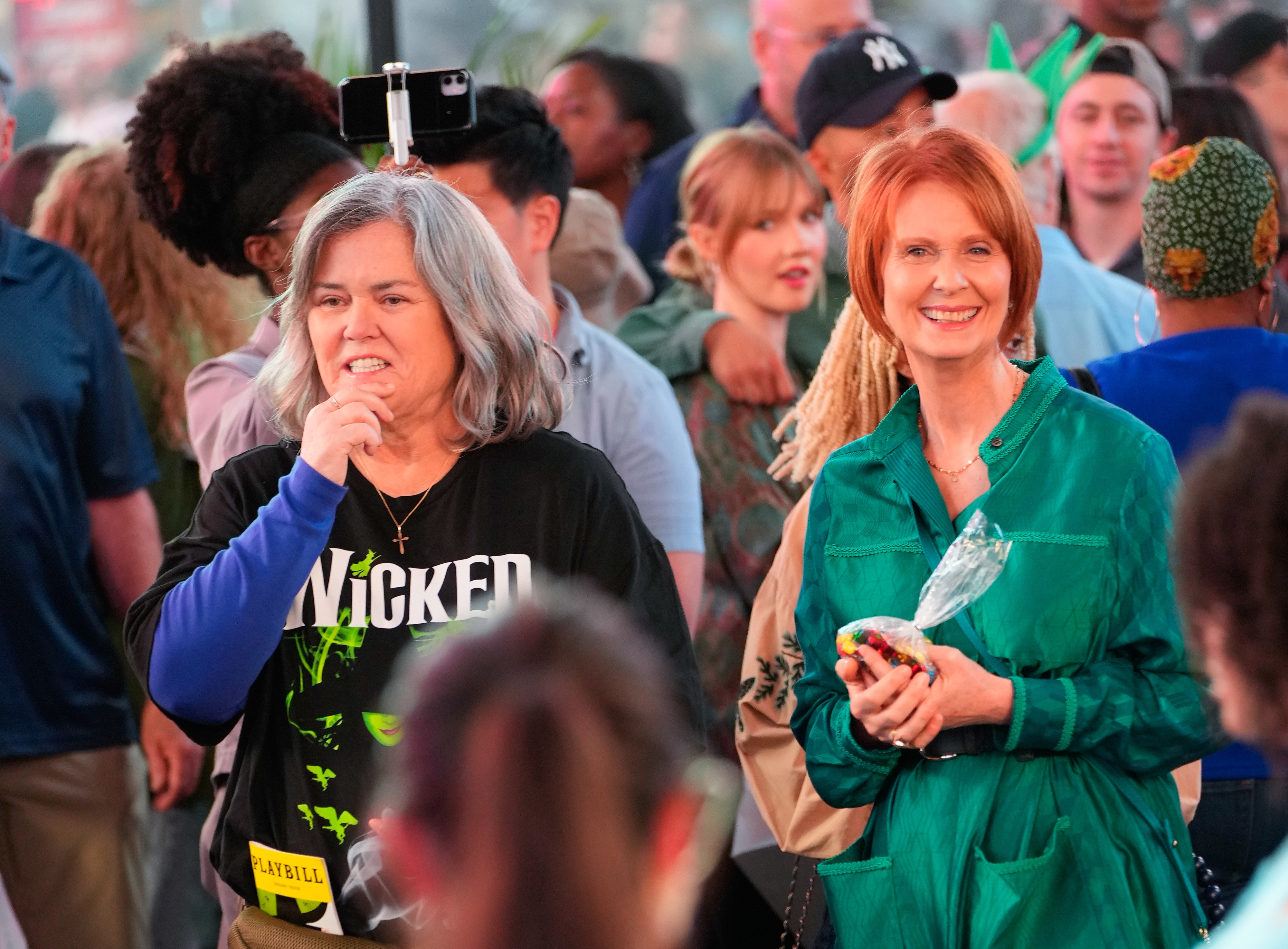 Rosie O&#x27;Donnell in a &#x27;Wicked&#x27; T-shirt and Cynthia Nixon in a green outfit are among a crowd of people, smiling at an event