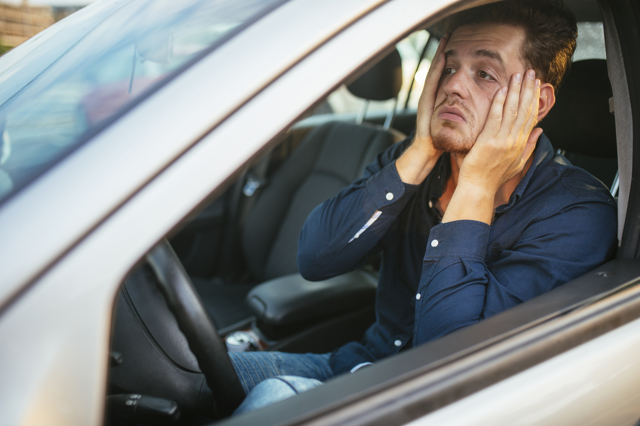 A man sits in a car, looking frustrated and resting his face in his hands. The image relates to an article about work and money