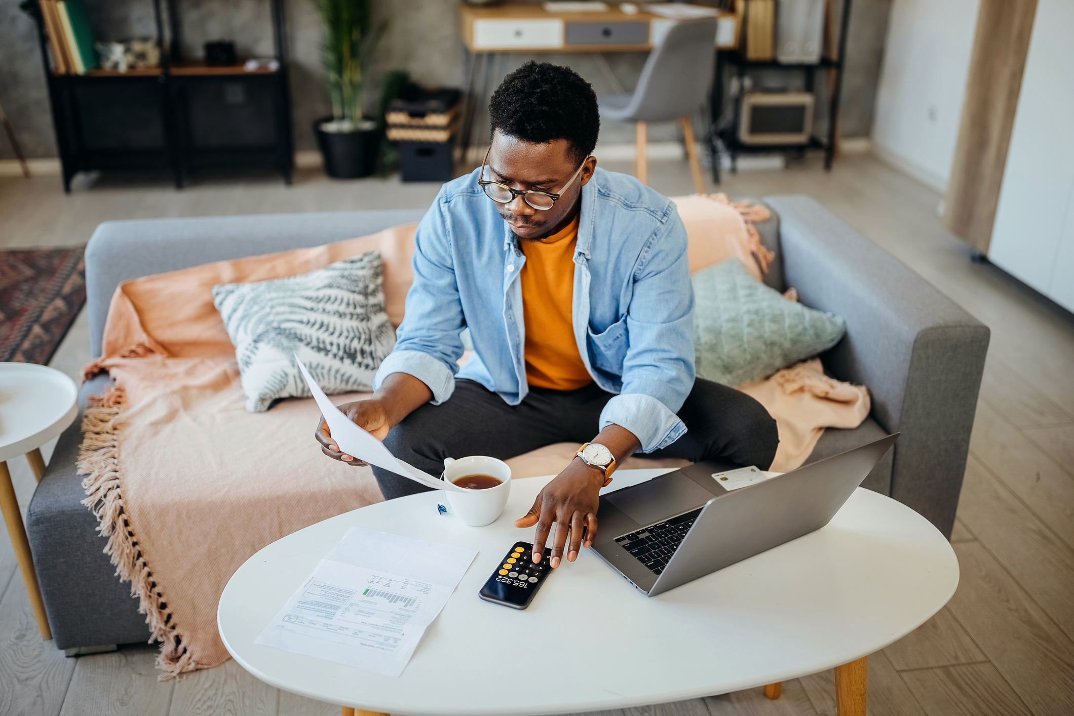 A person sits on a sofa, holding a document, with a laptop open, a cup of coffee, and financial papers on a white table in front of them, likely working from home
